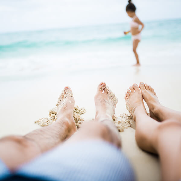 feet of couple relaxing on beach, their kid is playing at the edge of the water in front of them